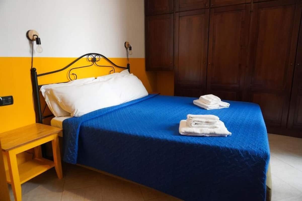 Un lit avec une couvre-lit bleue et des oreillers blancs, une table de chevet en bois et une armoire en bois à l’arrière-plan.