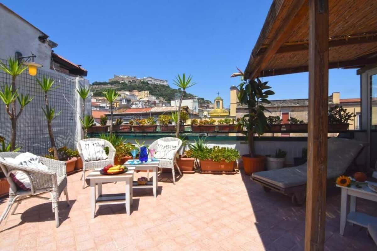 Une terrasse sur le toit, aménagée avec des meubles en osier, des plantes en pot et offrant une vue sur le paysage urbain qui s’étend sur une colline.