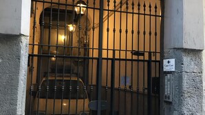 A black metal gate with a decorative lattice design, a sign reading "Case Cosi," and a small ventilation unit on the wall. - Case Così Chiaia (Naples)