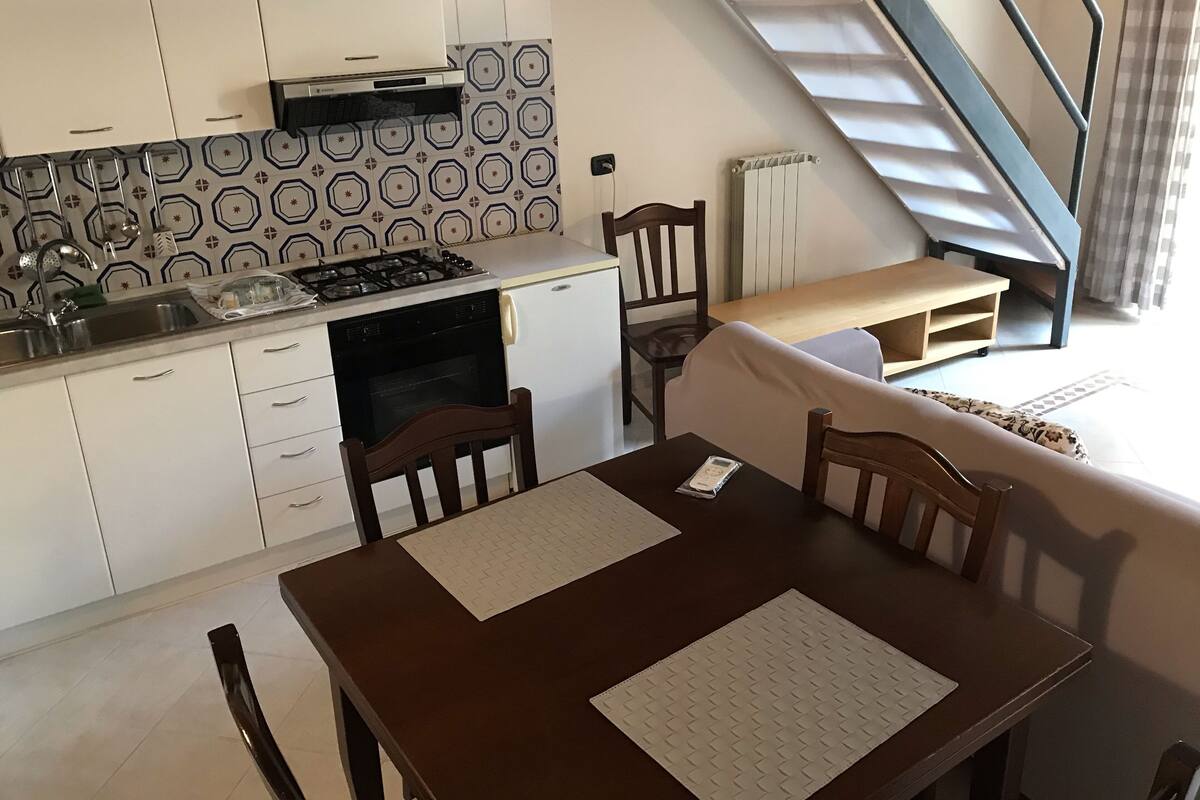Une cuisine compacte équipée d’une cuisinière à gaz, d’un évier et de meubles blancs. Une table en bois avec deux chaises se trouve dans l’espace repas, et un escalier est visible au fond.