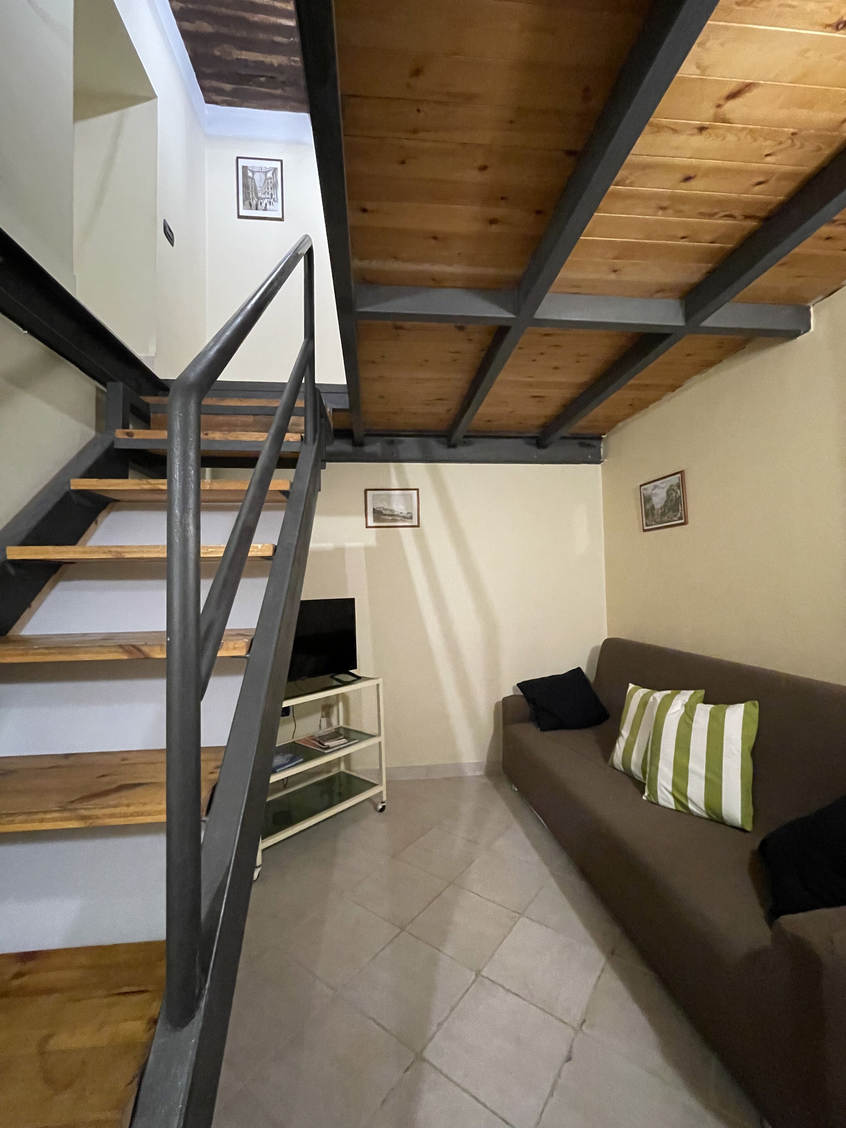 Una escalera con escalones de madera y barandillas de metal, un sofá marrón con almohadones a rayas y un televisor sobre un pequeño mueble.