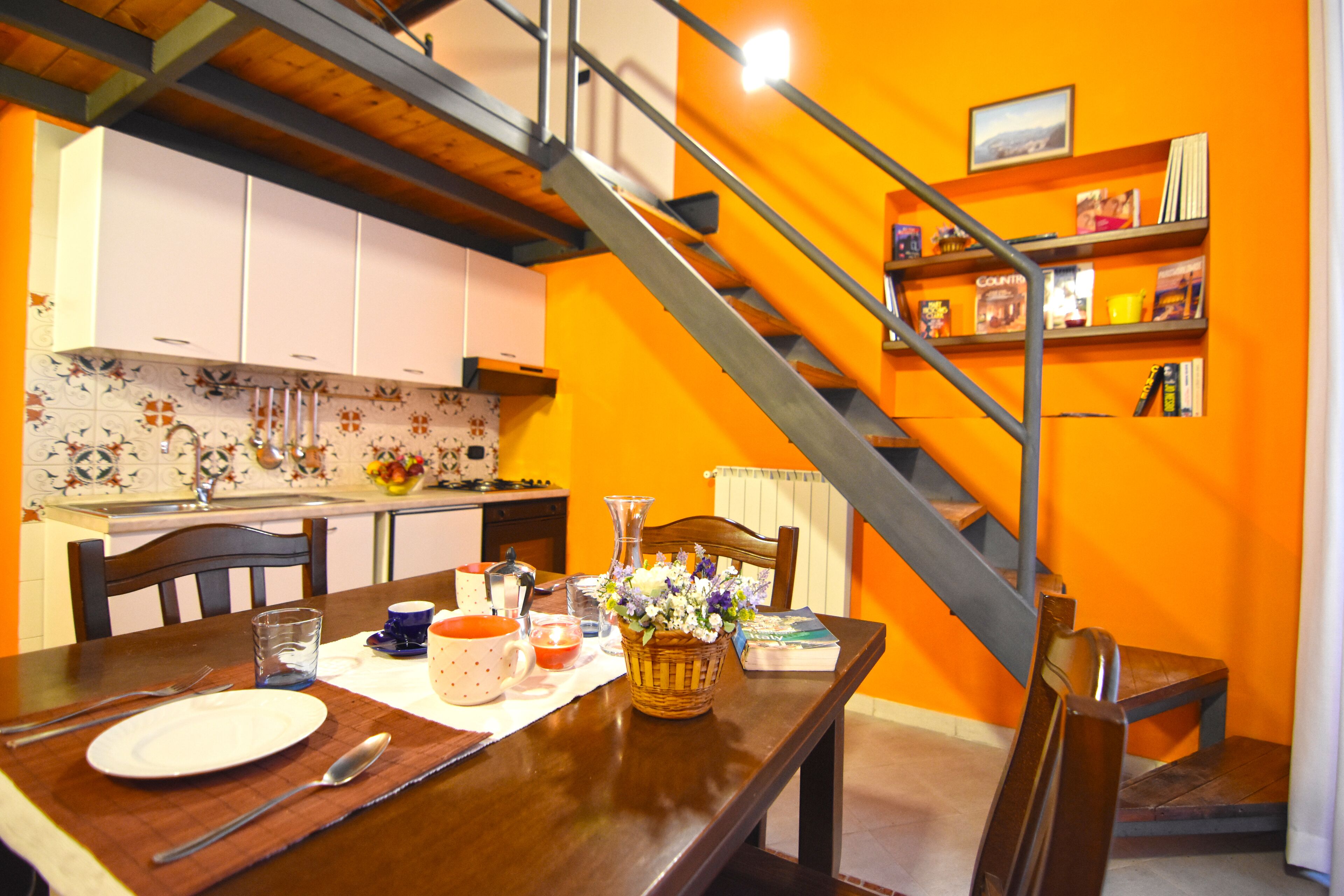 A dining area with a wooden table set for two, a kitchen in the background, and a staircase leading to an upper level.