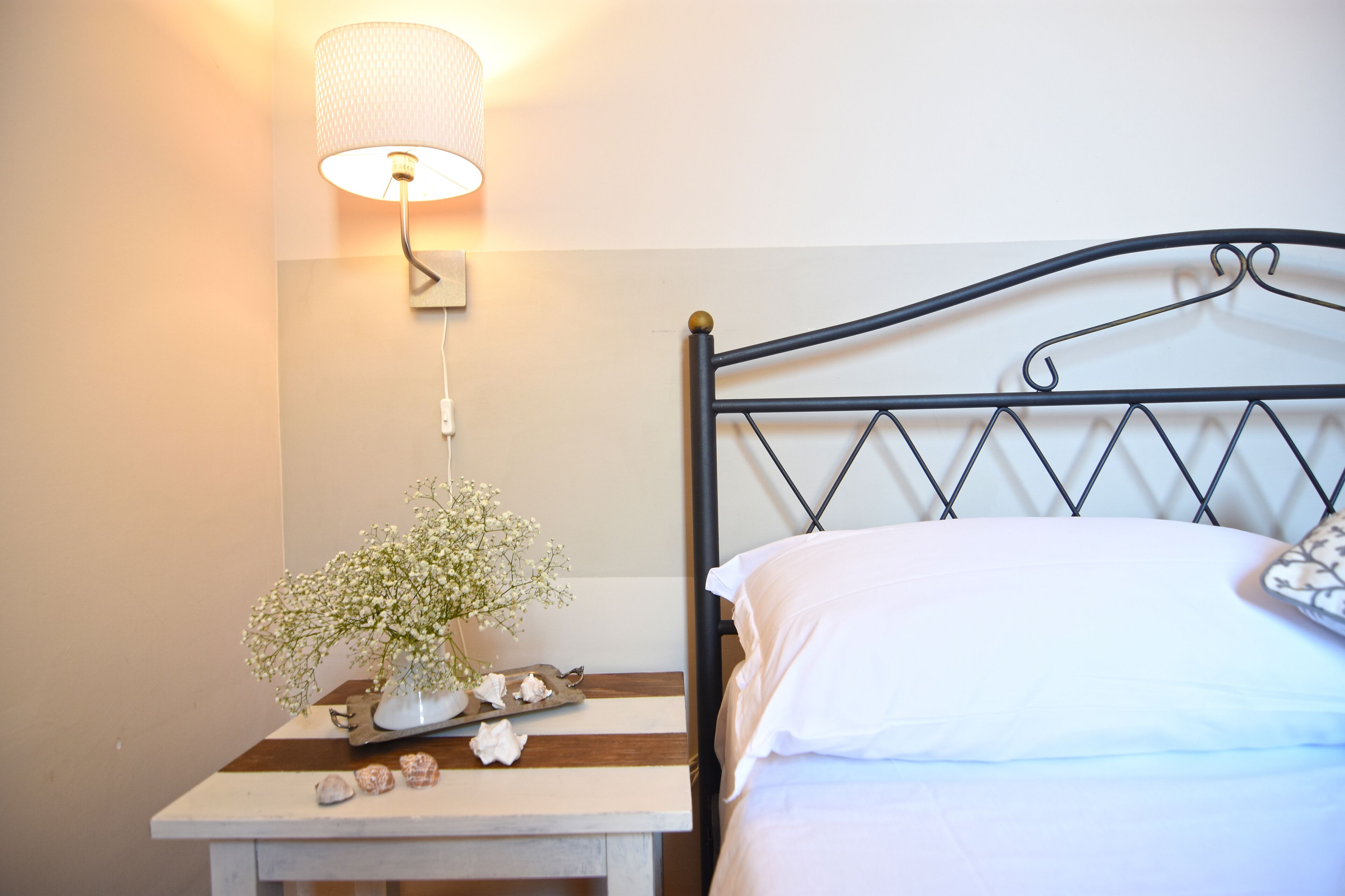 Un lit bien fait, avec du linge de lit blanc, une table de chevet avec une lampe, un vase rempli de fleurs et un plateau orné de coquillages.