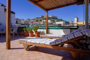 Un fauteuil de jardin en bois, doté d’un coussin à rayures, est placé sur une terrasse carrelée, d’où l’on peut admirer un paysage urbain avec ses bâtiments et une colline.