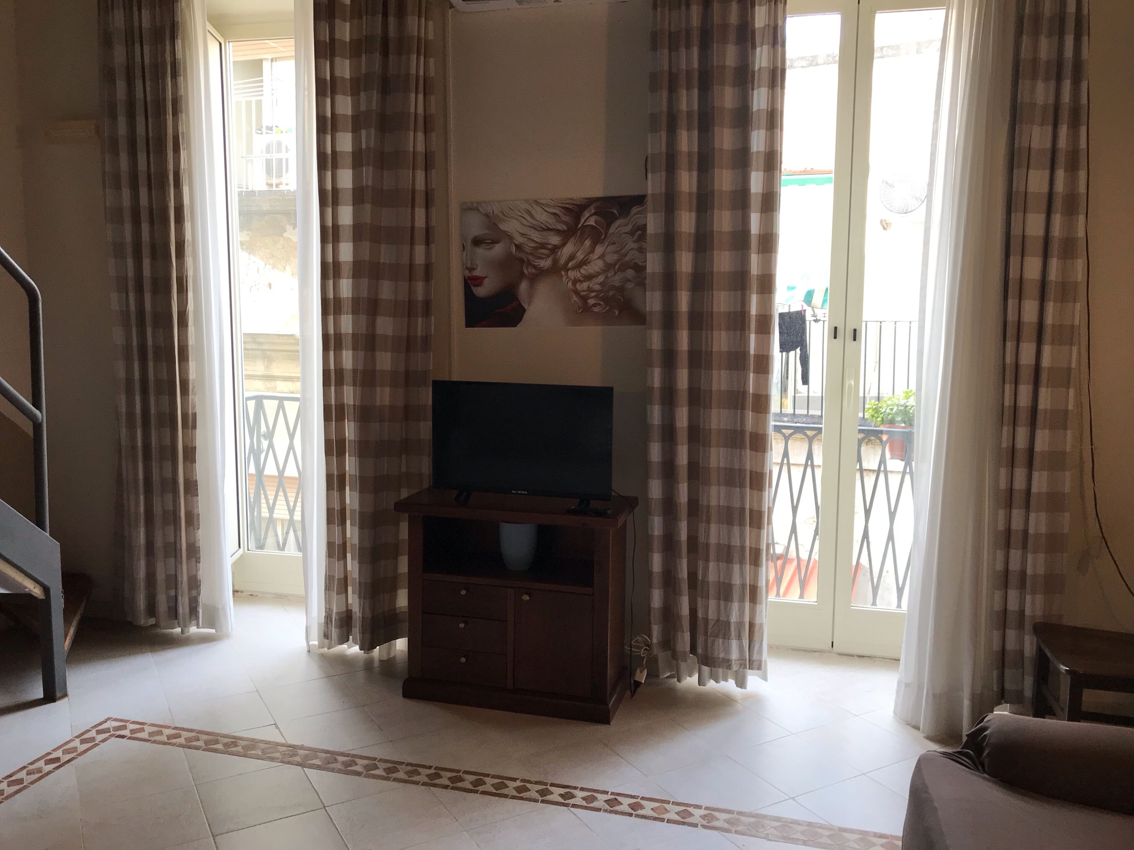 Un soggiorno con televisore su un mobile in legno, tende a quadri e pavimento piastrellato con motivo geometrico.
