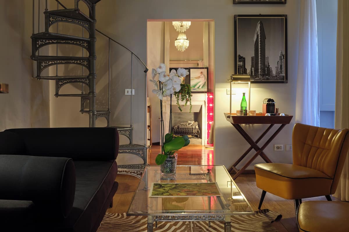 Una sala de estar con una escalera, una mesa de centro de vidrio, un sofá, un sillón y una araña.