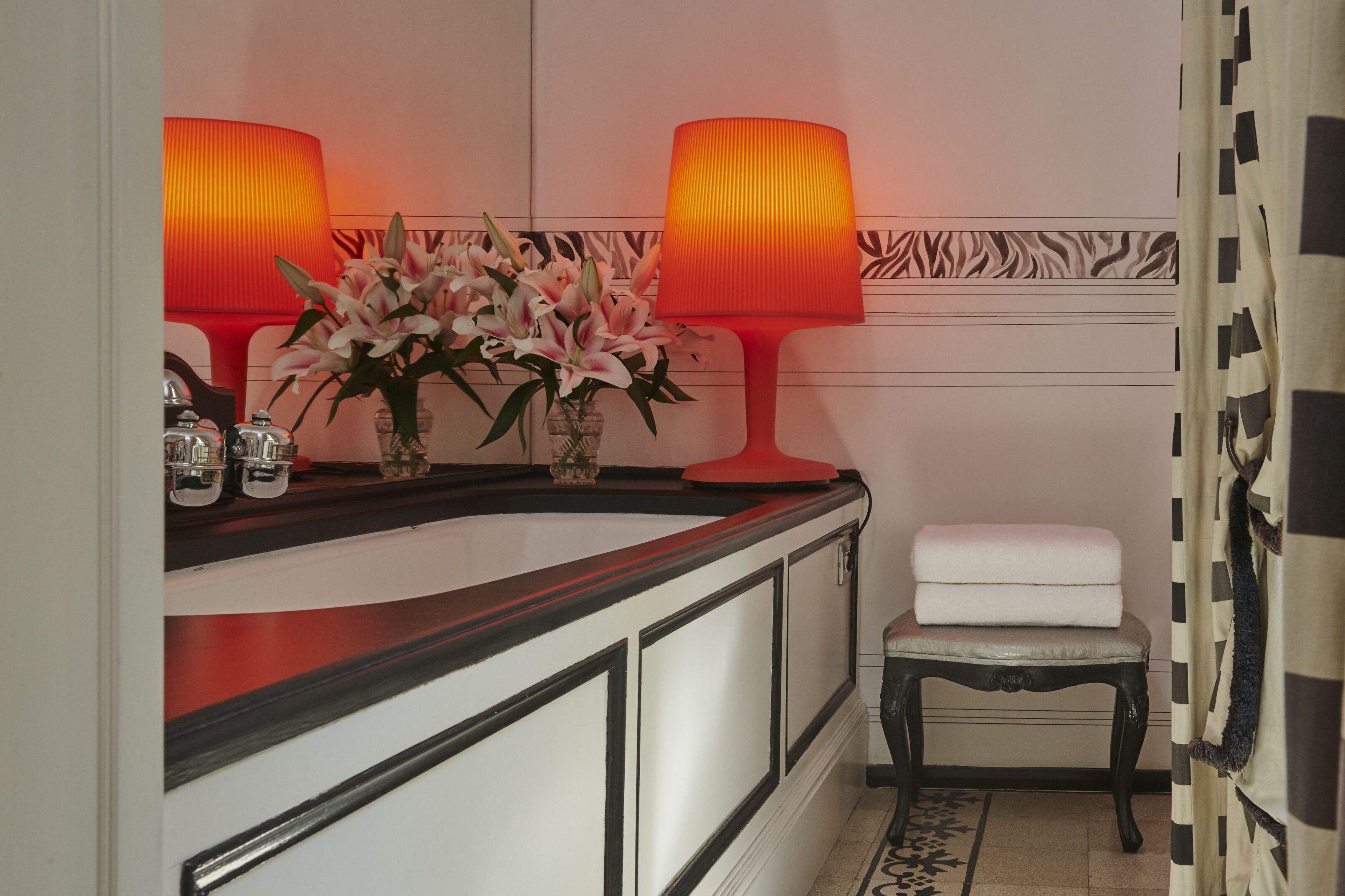 Une salle de bain avec un meuble-lavabo blanc, deux lampes rouges, un vase avec des fleurs roses, une serviette blanche posée sur une chaise et un mur à motifs.