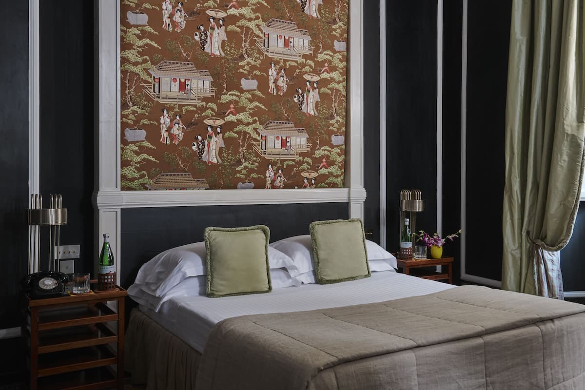 Habitación de hotel con una gran obra de arte tradicional en la pared, una cama bien hecha con dos almohadas, una mesita de noche con una lámpara, una botella y un vaso.
