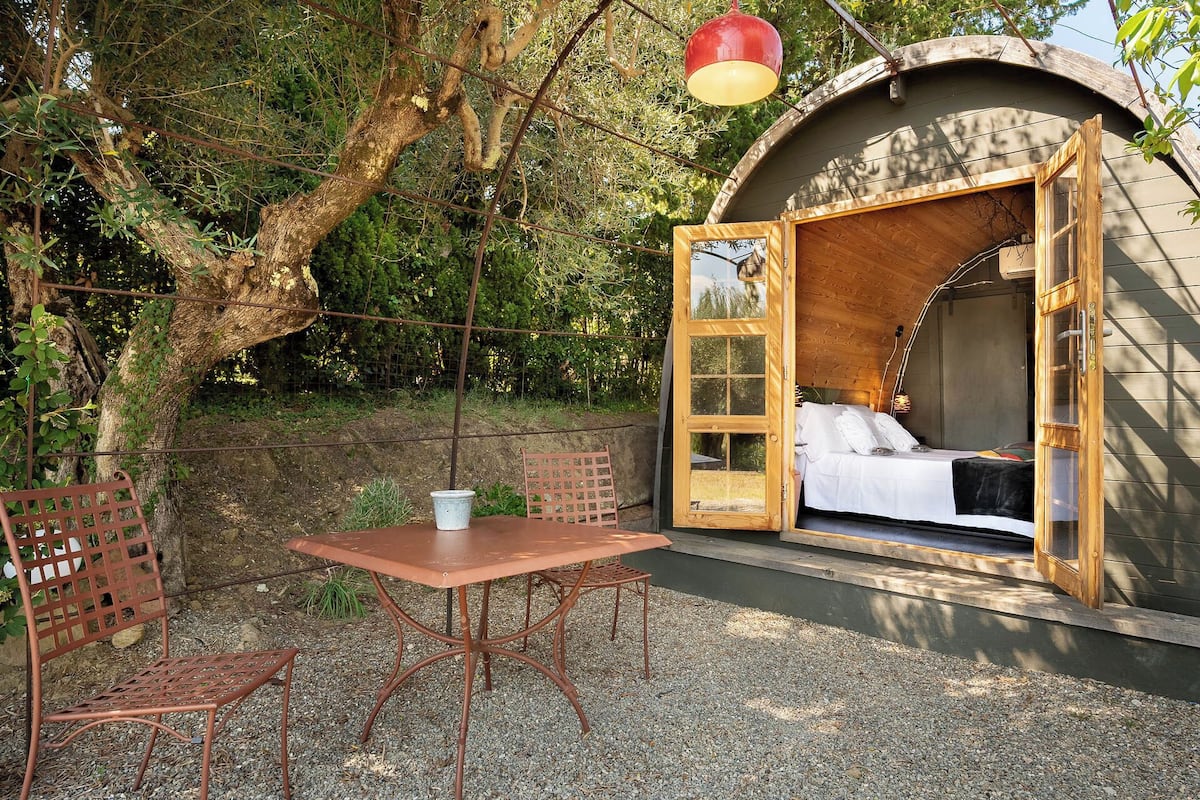 Un espacio al aire libre acogedor con una cama, una mesa y sillas, rodeado de árboles.