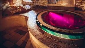 Una habitación con paredes de piedra, un banco de piedra, una bañera con luz rosa y una ventana con vistas.