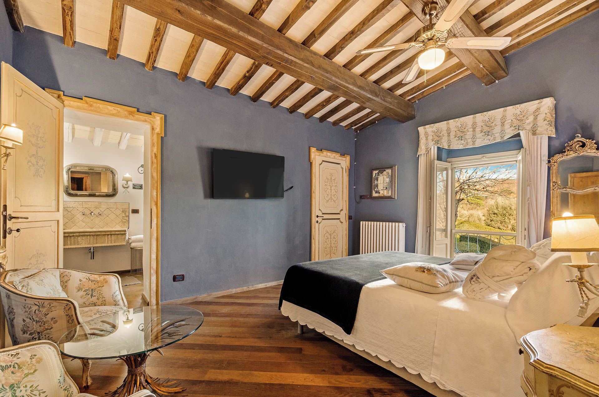 A bedroom with a bed, a sitting area with a glass table, a television, and a window with a view.