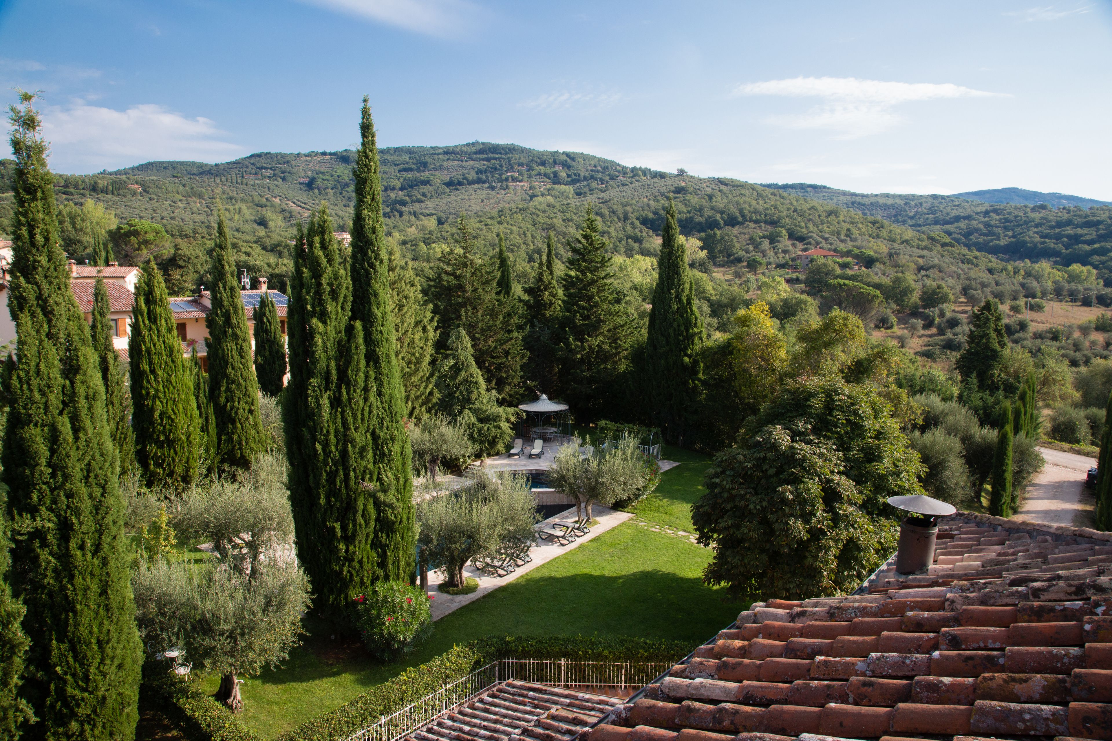 A courtyard with a gazebo, surrounded by tall cypress trees and a view of a hilly landscape.