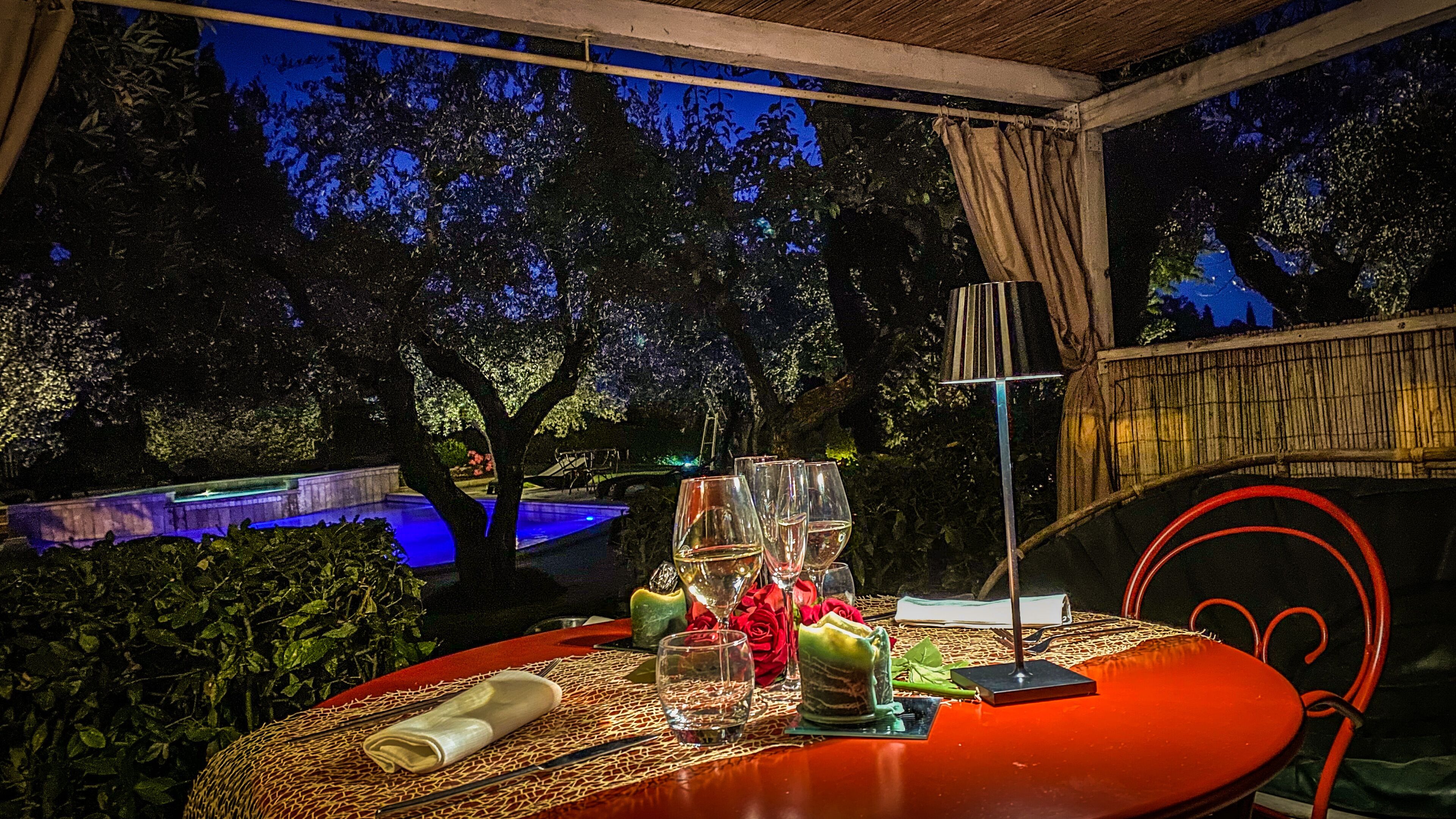 An outdoor dining area with a table set for four, surrounded by lush greenery and a pool in the background.