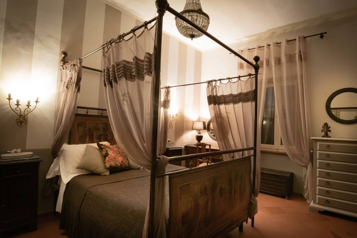 Una cama con dosel y cortinas translúcidas, una mesita de noche con lámpara, un aparador y un espejo en la pared.