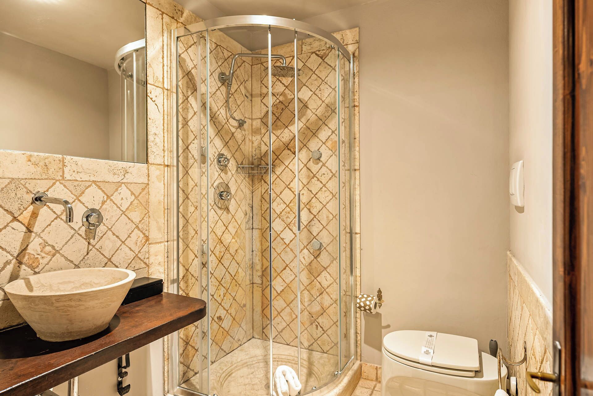 Une salle de bain dotée d’une douche à parois de verre, d’un mur recouvert de carreaux de marbre, d’un meuble-lavabo en bois avec un lavabo en forme de coupe et de toilettes.