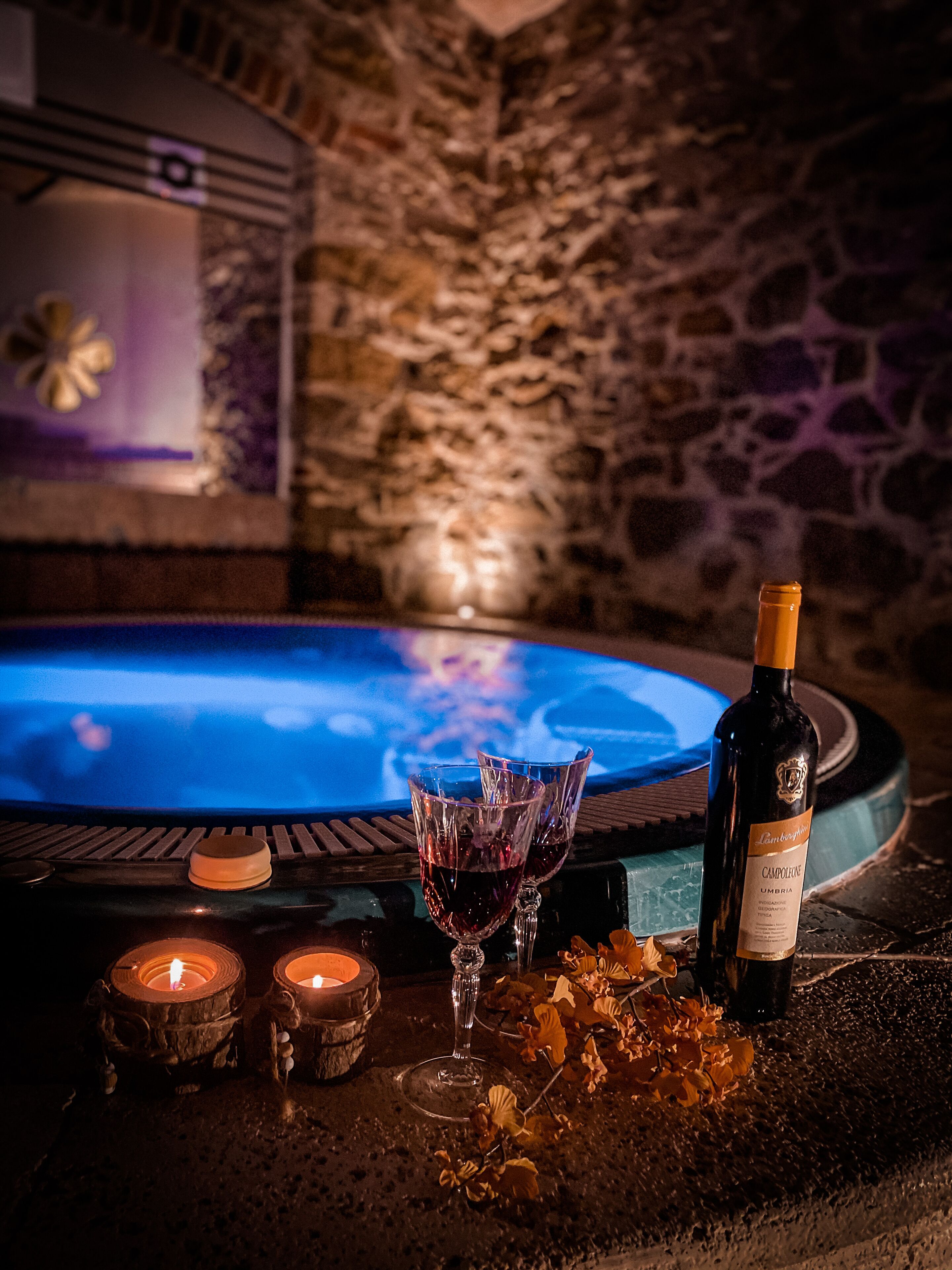 Un espace spa avec un mur en pierre, un jacuzzi, des bougies et une bouteille de vin posée sur une table.