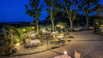 Un área de comedor al aire libre con mesas y sillas, rodeada de árboles y suelo de piedra.