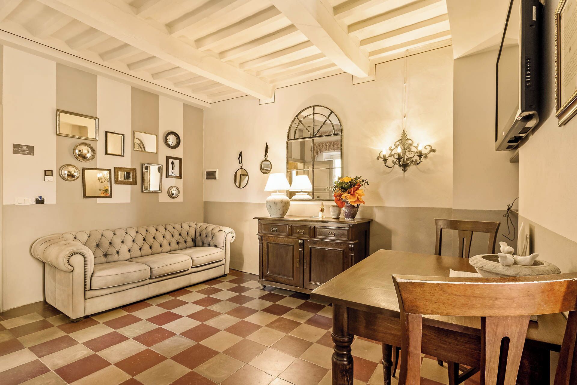 A dining area with a wooden table and chairs, a checkered floor, a sofa, and a wall with framed mirrors and a chandelier.
