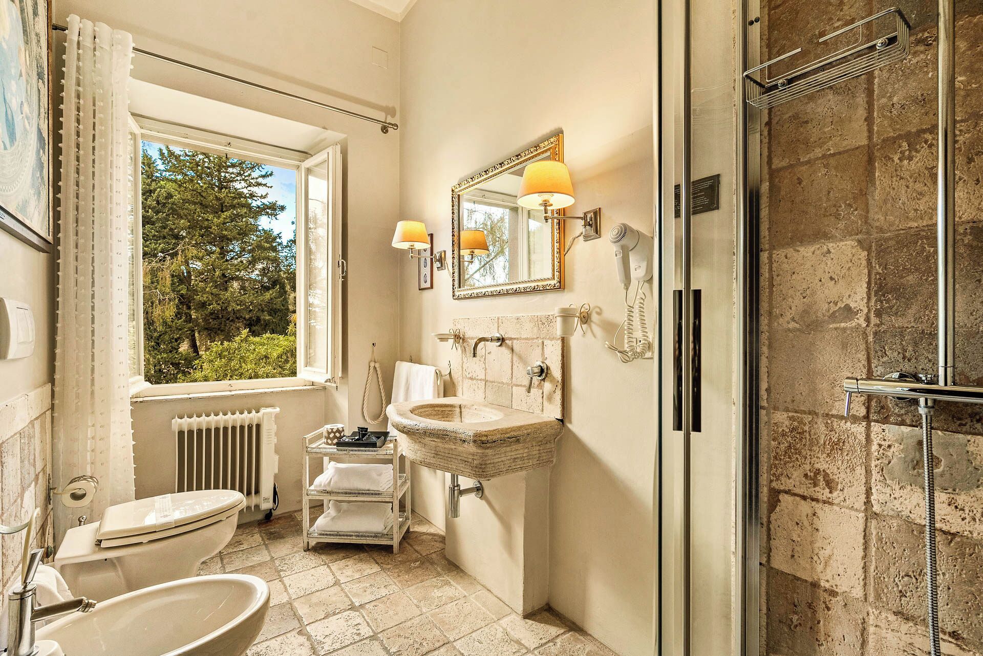 A bathroom with a large window, a stone wall, a sink, a toilet, and a shower.