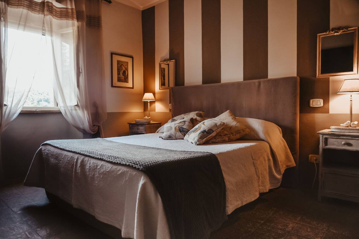 Una cama bien tendida con cabecera, una mesita de noche con lámpara y una ventana con cortinas de encaje.