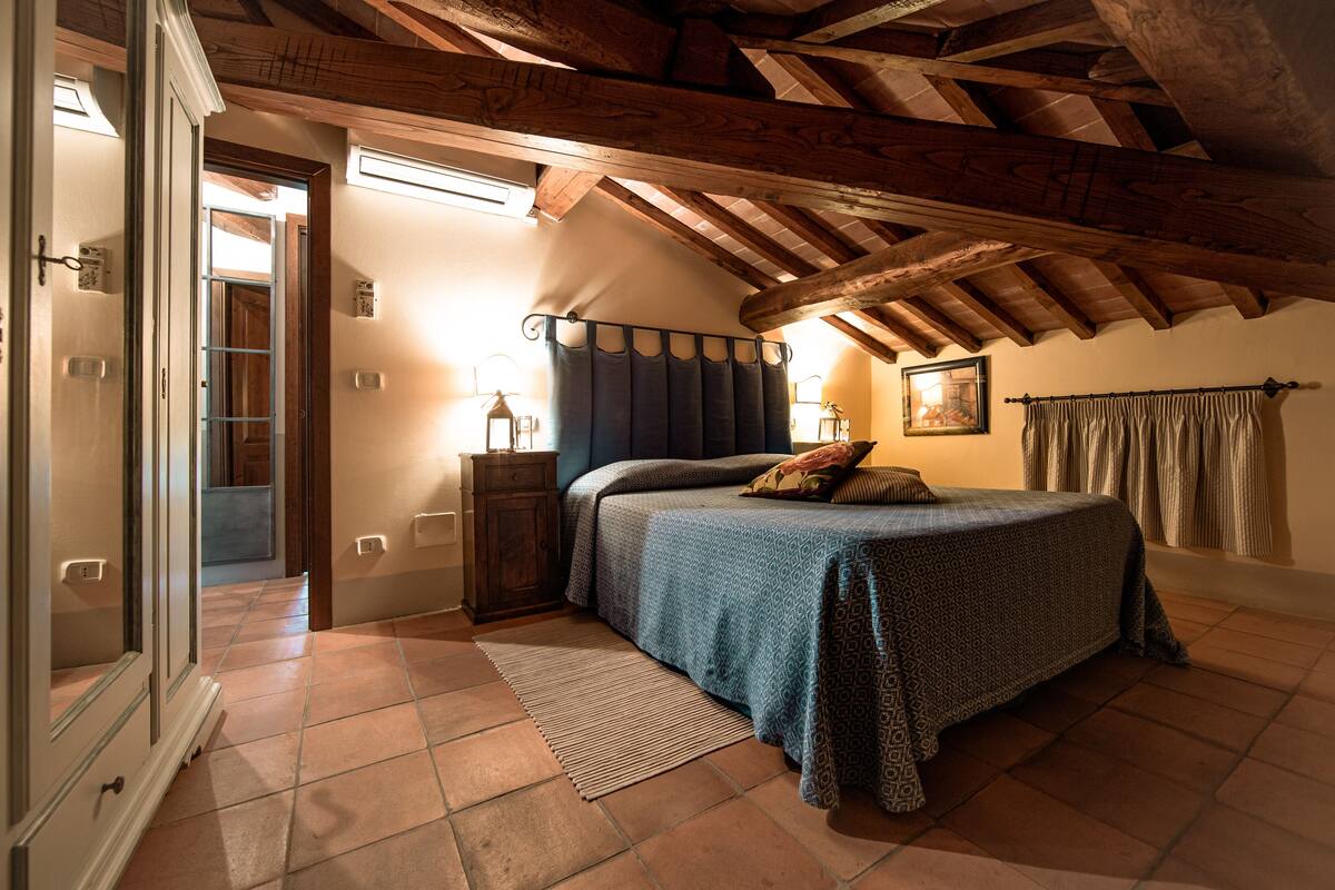 Un dormitorio con techo inclinado, una cama con colcha estampada, una mesita de noche con lámpara y vistas al exterior a través de una puerta.