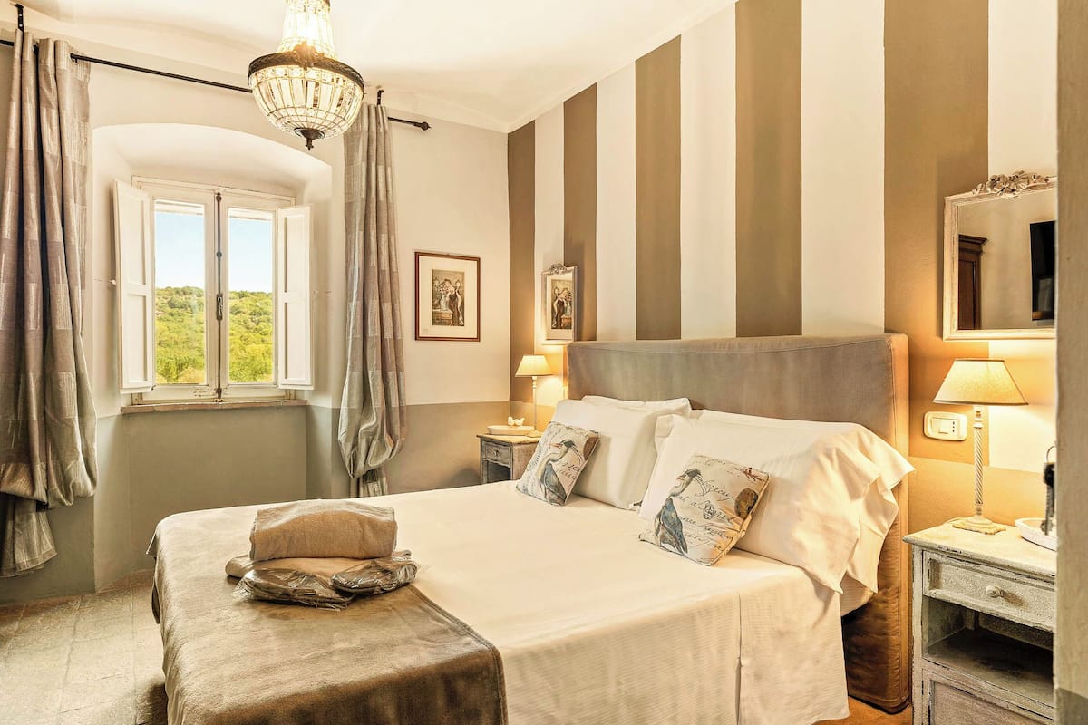 Un dormitorio con una cama grande, una mesita de noche, una lámpara, una ventana con cortinas y un candelabro.