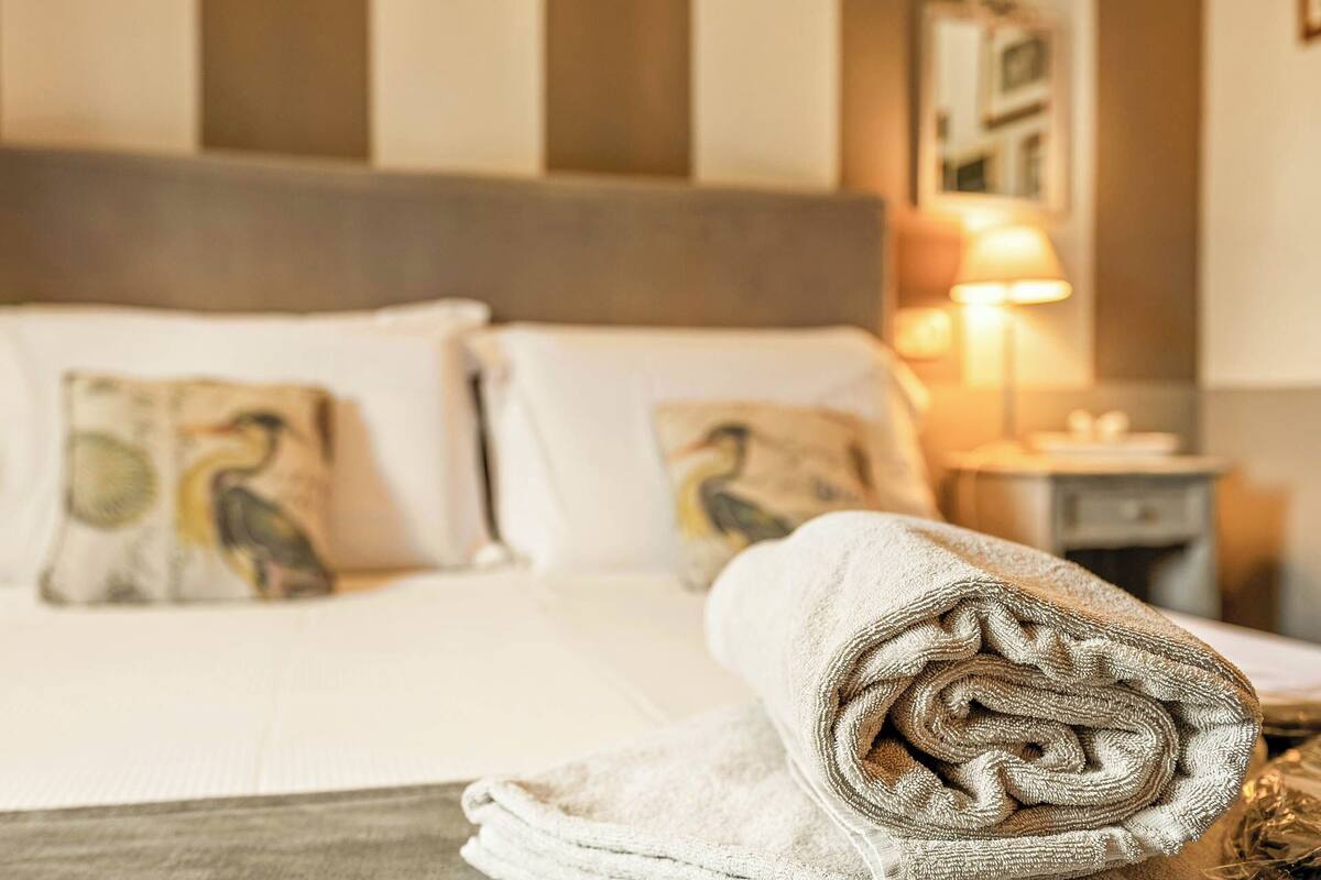 Una cama bien hecha con sábanas blancas y dos almohadas con diseños de pájaros, una toalla enrollada y una mesita de noche con una lámpara.