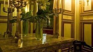 A room with a large mirror reflecting a chandelier, a candelabra, and a vase with flowers. There is a bench and a table with a vase of flowers. - Palazzo Vecchio (Florence)