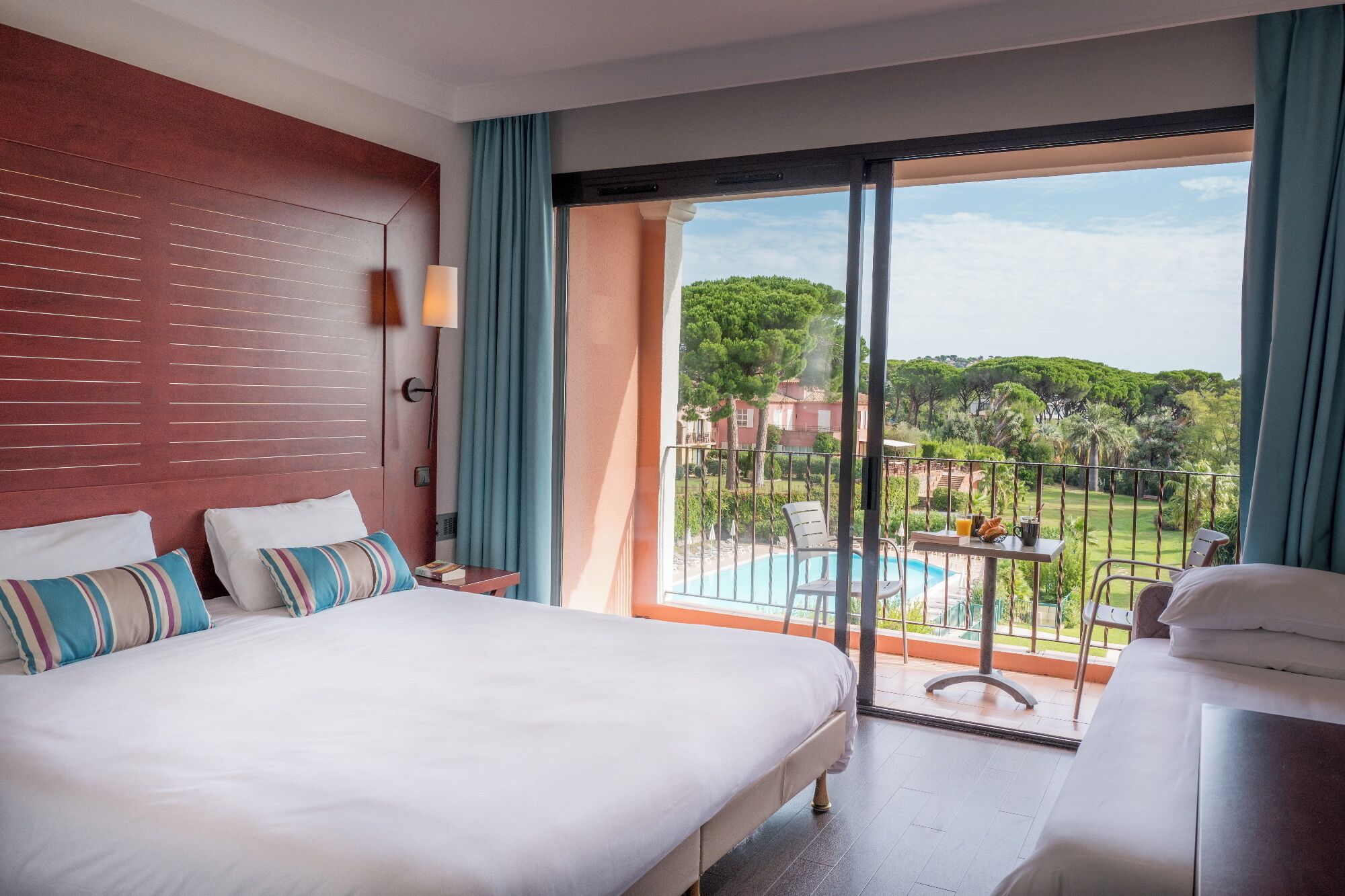 Habitación de hotel con cama, una mesita con una bebida, una silla y vistas a una piscina y zonas verdes.