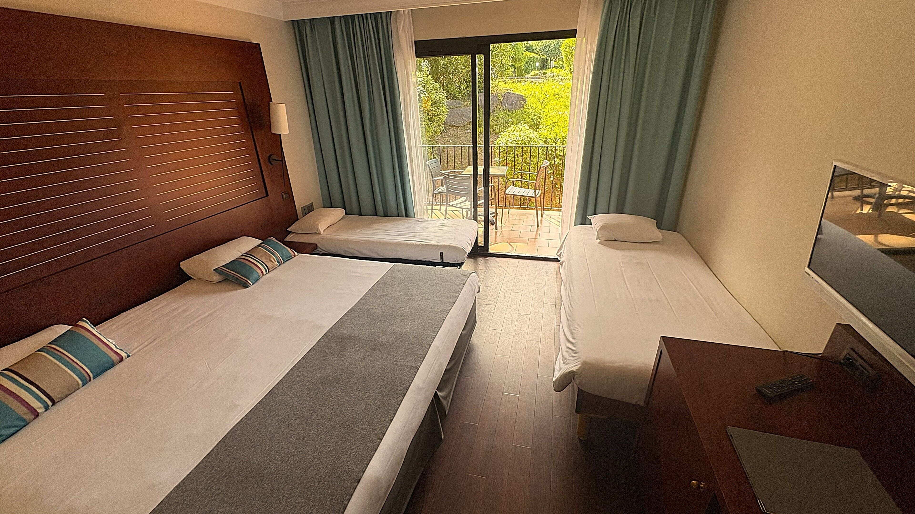 A hotel room with two beds, a wooden headboard, a small desk with a TV, and a balcony with a view of greenery.