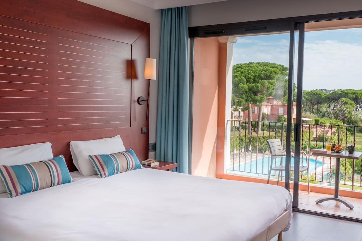 A hotel room with a large bed, a nightstand, a lamp, a view of a swimming pool, and a balcony.