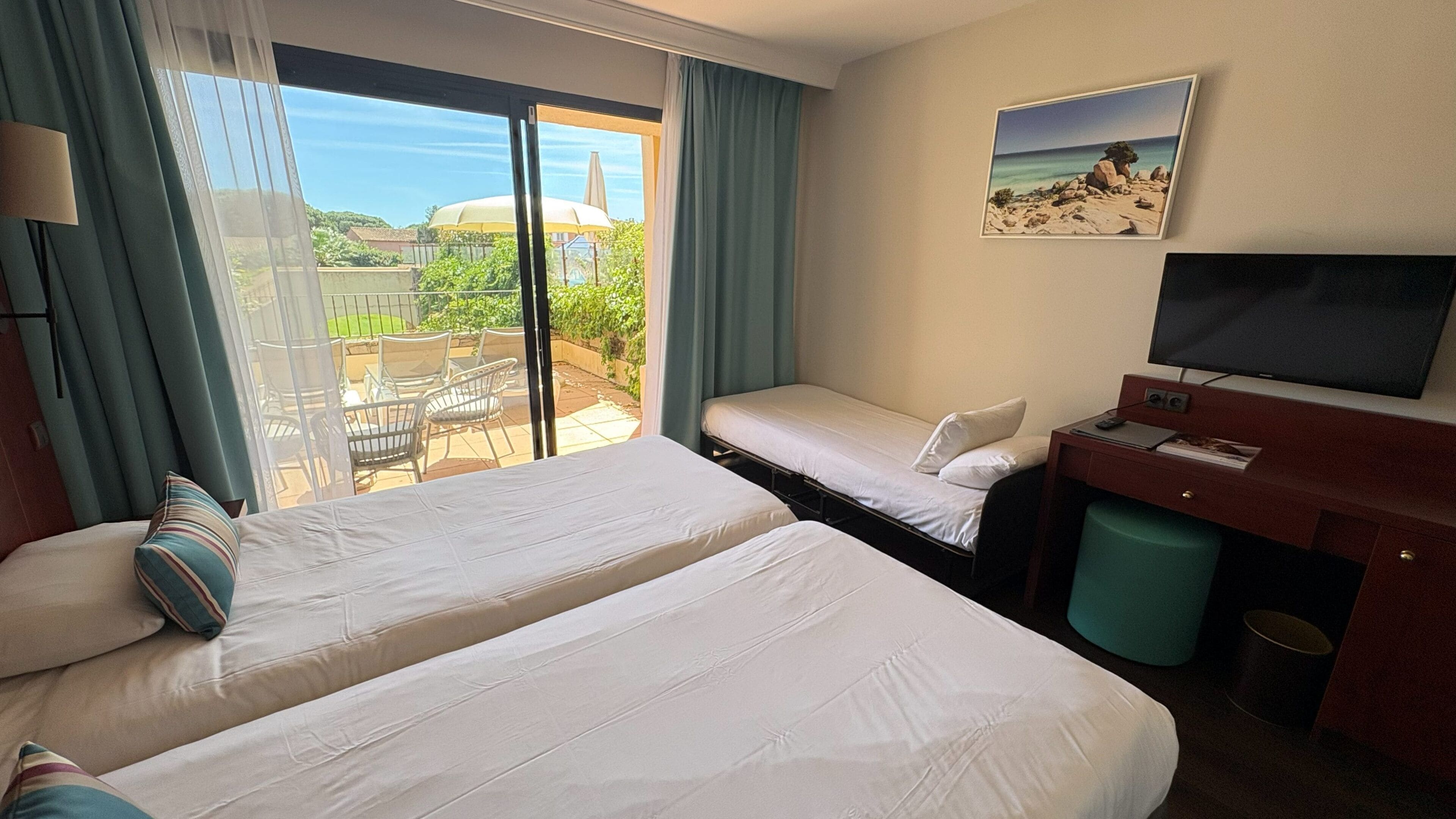 Ein Hotelzimmer mit zwei Betten, einem Schreibtisch mit Fernseher und einem Balkon mit Gartenmöbeln.