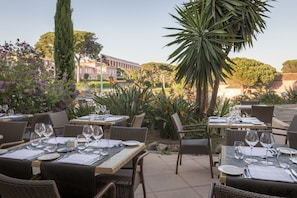 Un área de comedor al aire libre con mesas dispuestas para dos, rodeada de follaje y vistas a edificios al fondo.