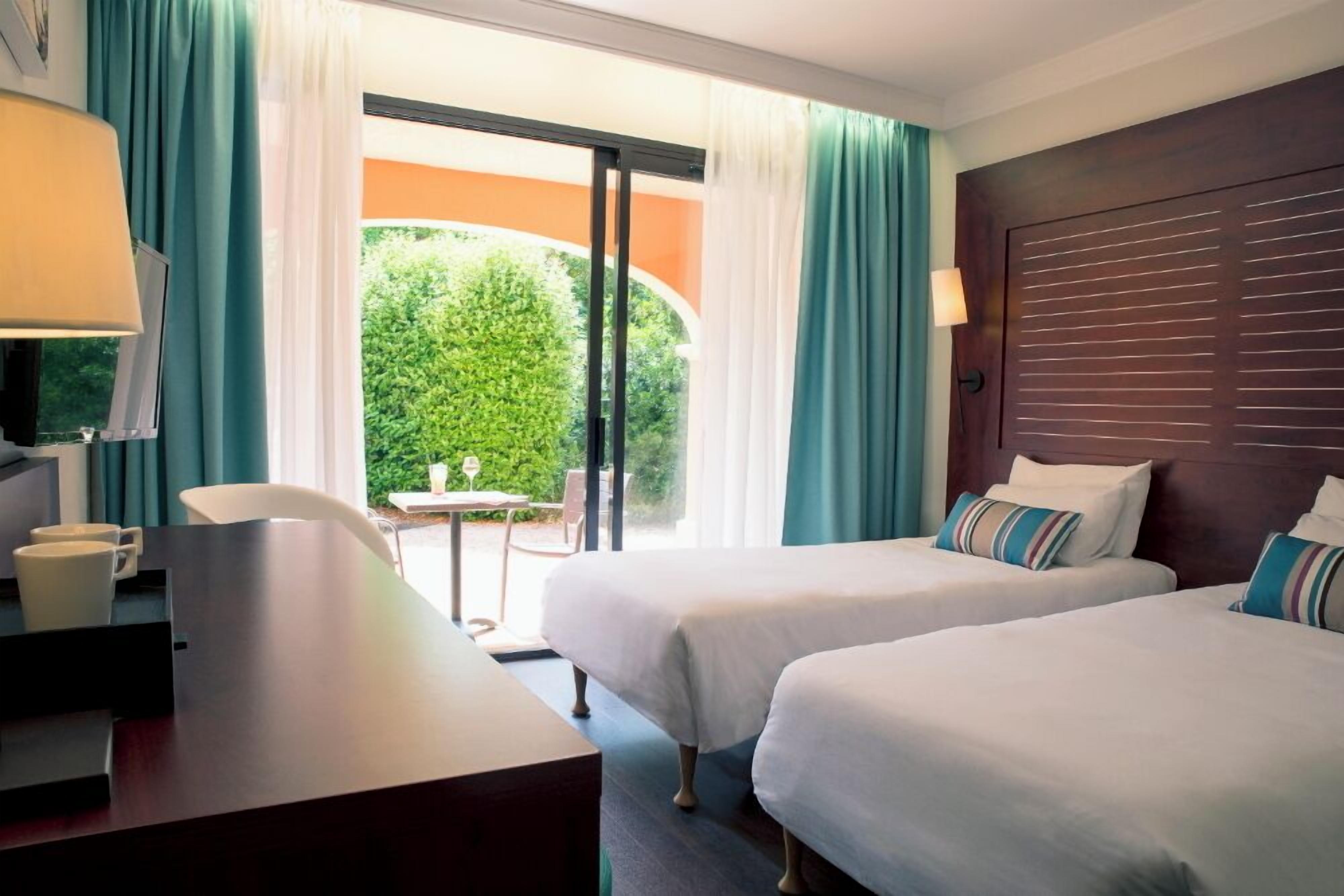 A hotel room with two beds, a desk, a chair, and a view of greenery through the window.