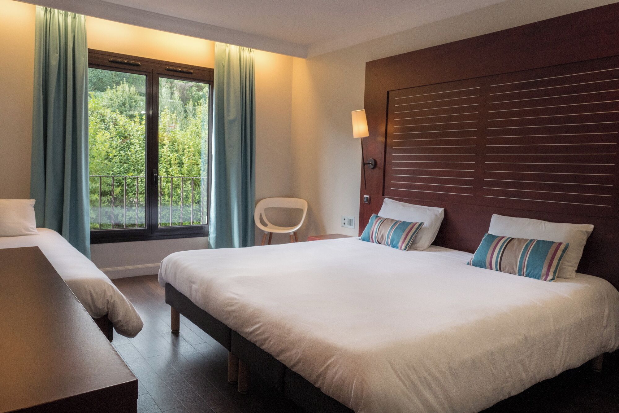 A hotel room with a bed, a chair, a window with curtains, and a view of greenery.