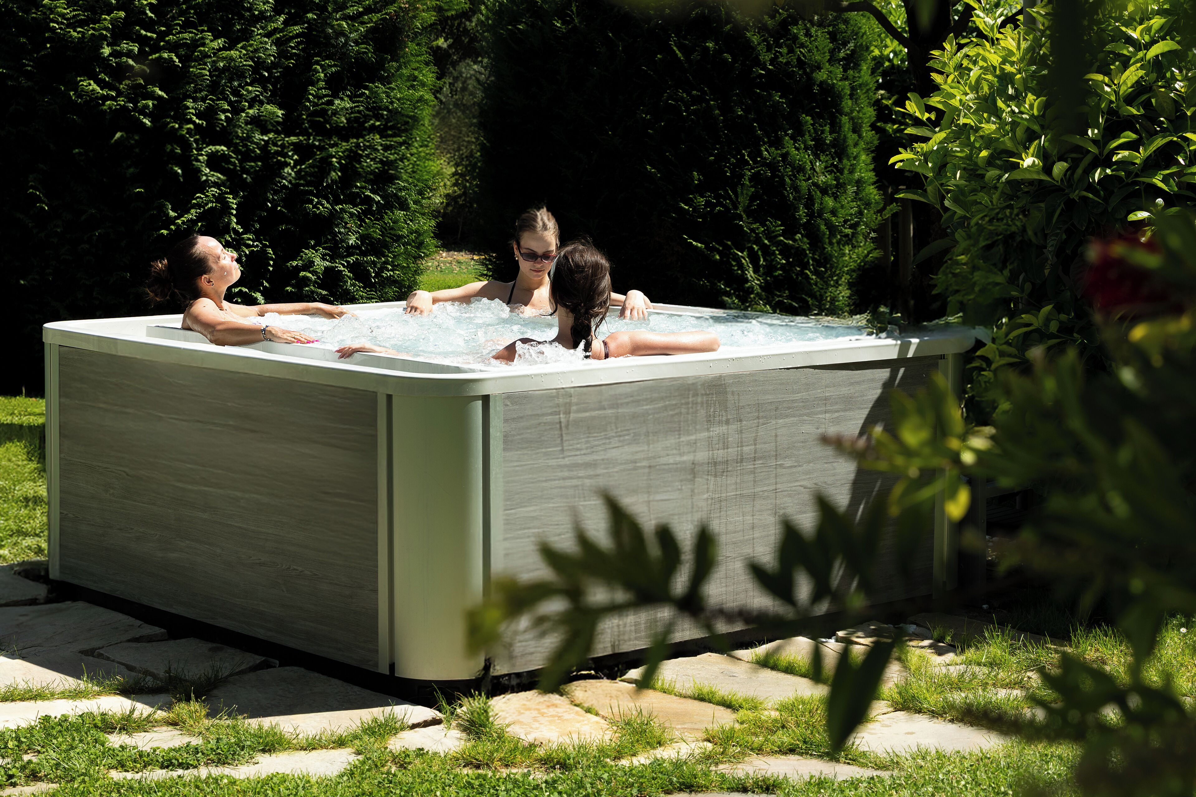 Un bain à remous rectangulaire à la surface lisse, entouré d’un chemin de pierre et d’une végétation luxuriante.
