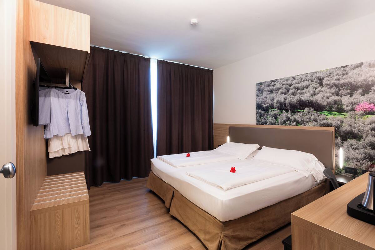 Une chambre d’hôtel avec un lit en bois, une armoire, un bureau et une grande fenêtre avec des rideaux.