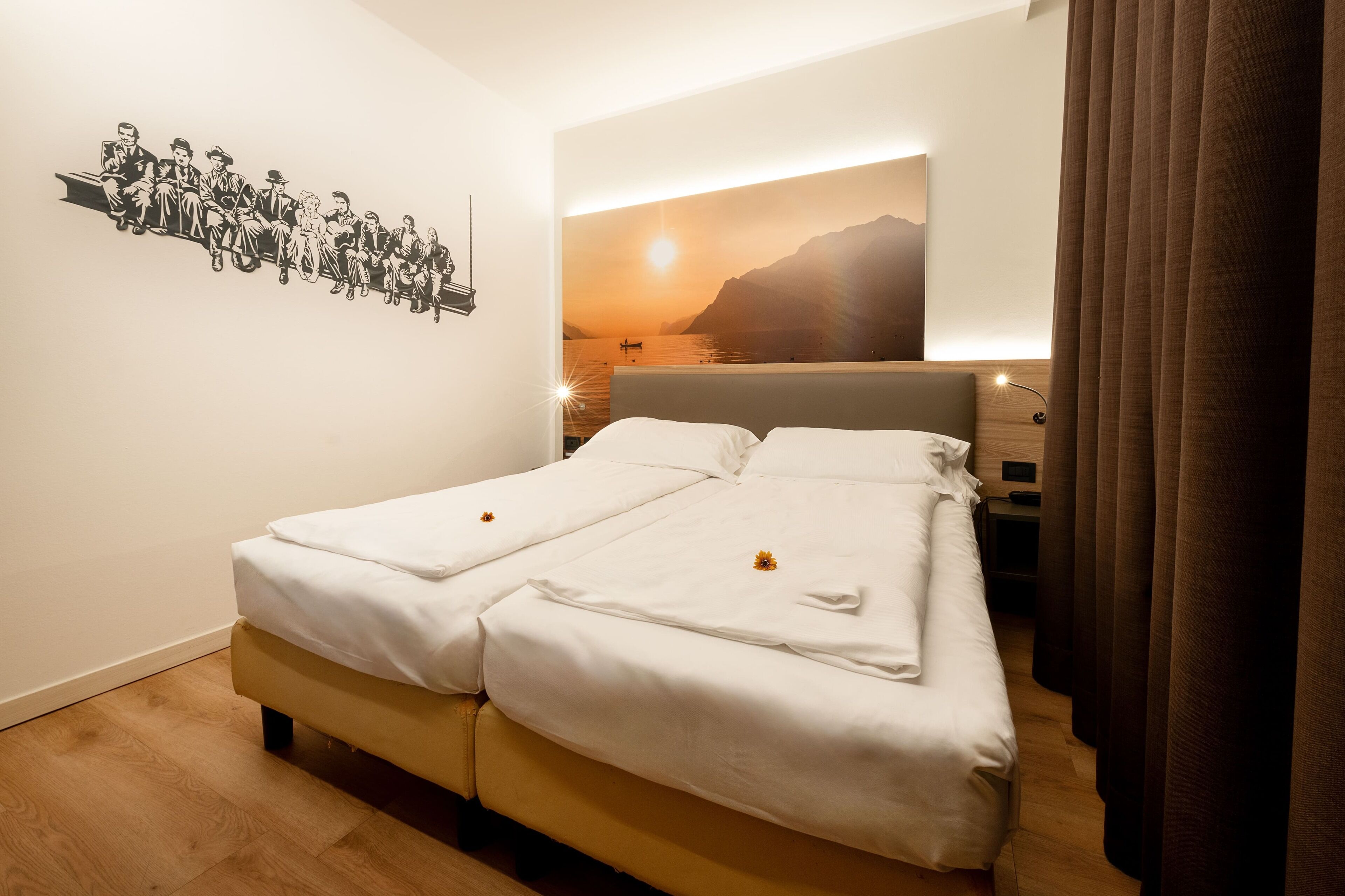 Een tweepersoonsbed met witte lakens, een kunstwerk aan de muur waarop een groep mensen te zien is, en uitzicht op een zonsondergang door een raam.