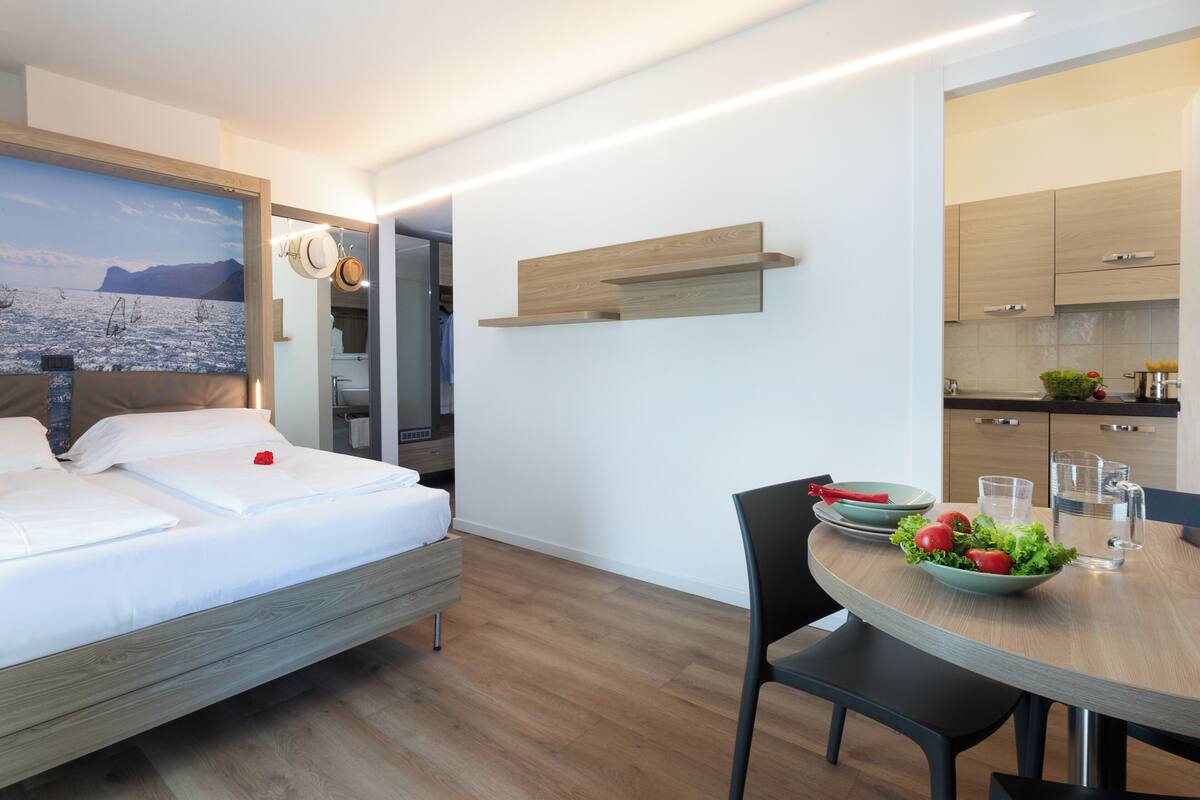 Une chambre d’hôtel moderne comprenant un lit, une table à manger avec des chaises, un coin cuisine et une salle de bain visible à travers une porte ouverte.
