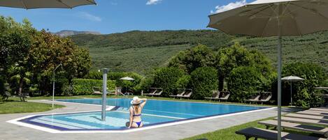 A swimming pool with lounge chairs and umbrellas, surrounded by greenery and hills.