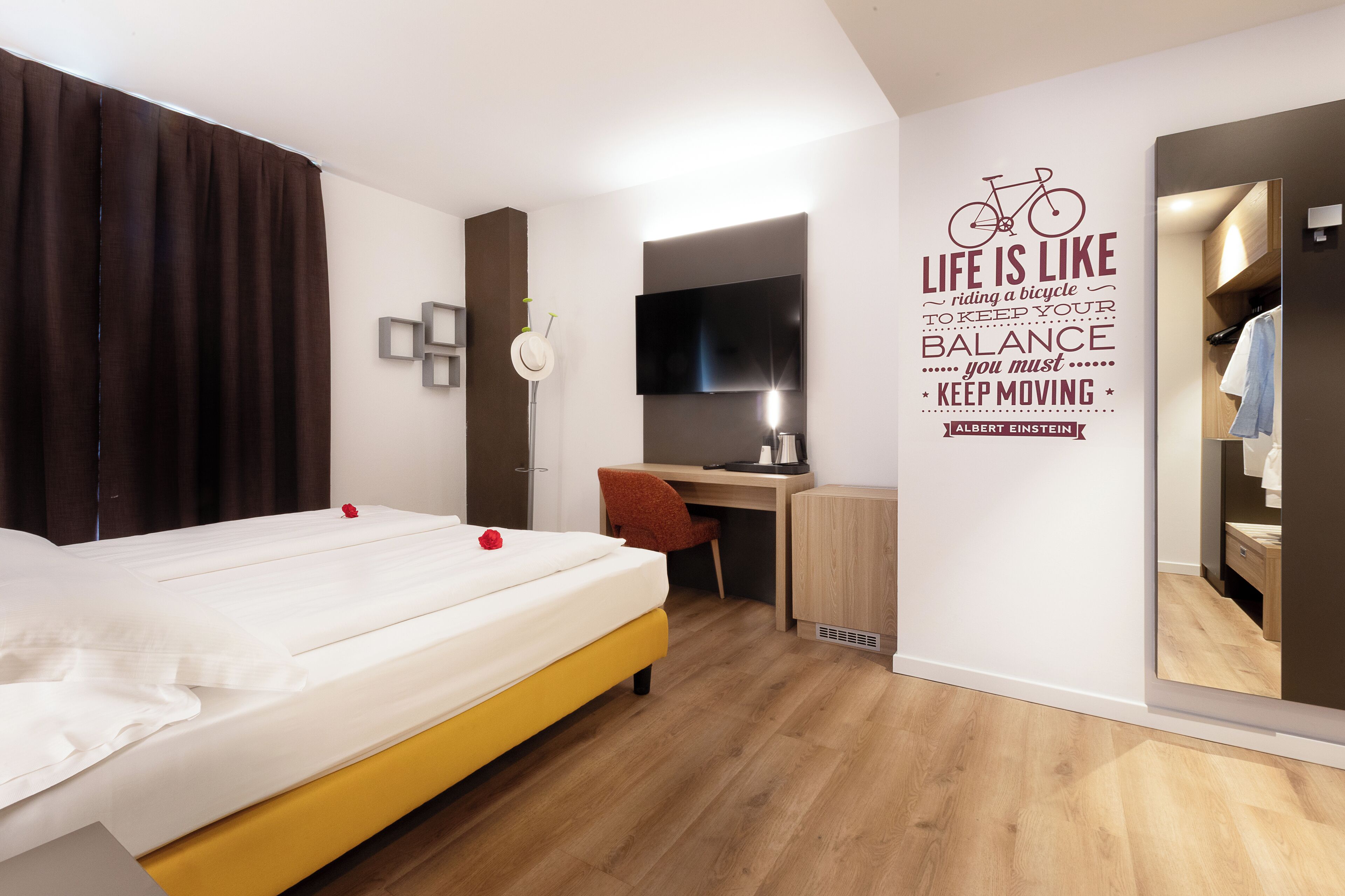 A modern hotel room with a bed, a desk, a chair, a TV, and a motivational quote on the wall.