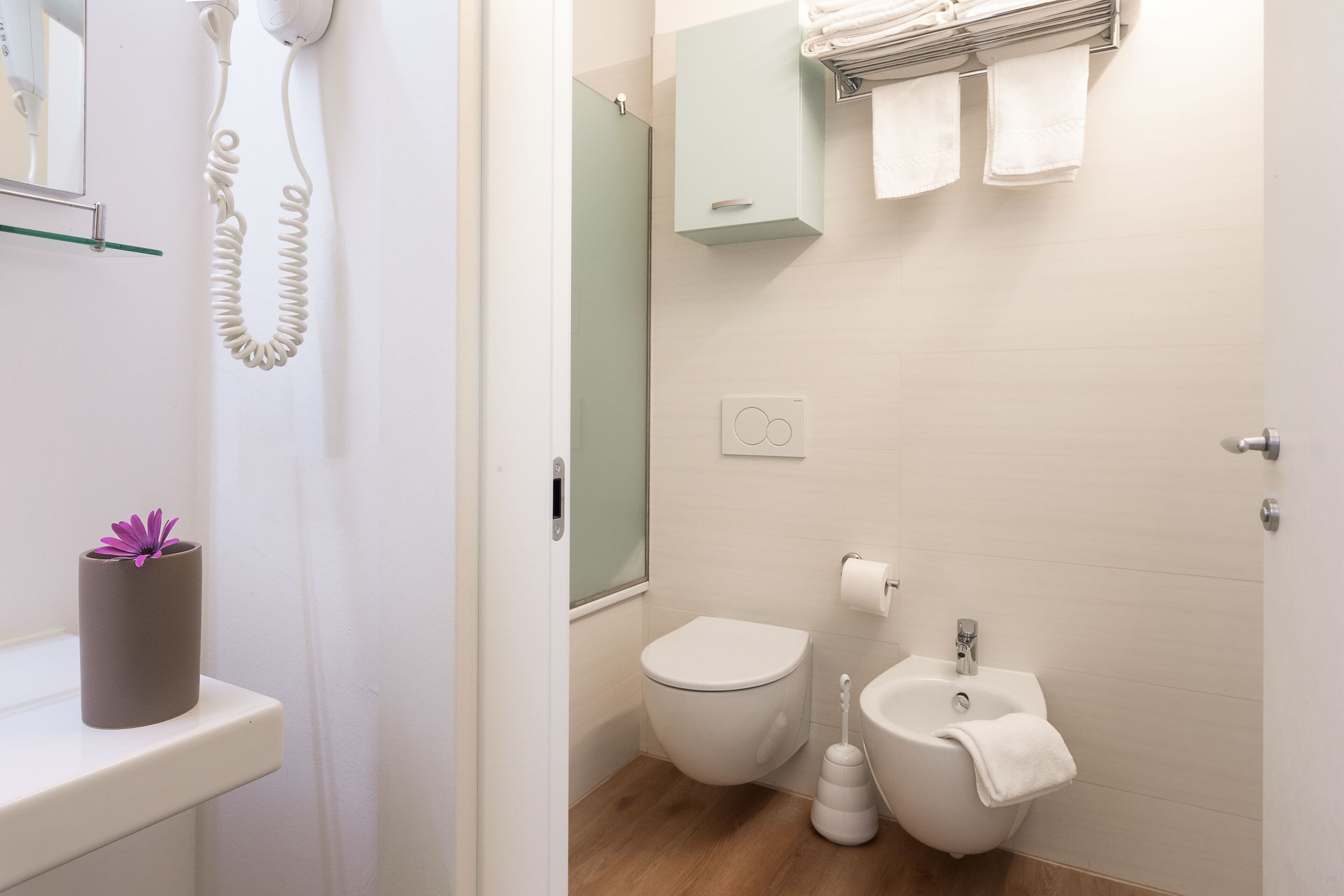 A bathroom with a toilet, bidet, and a glass shelf with a flower vase.