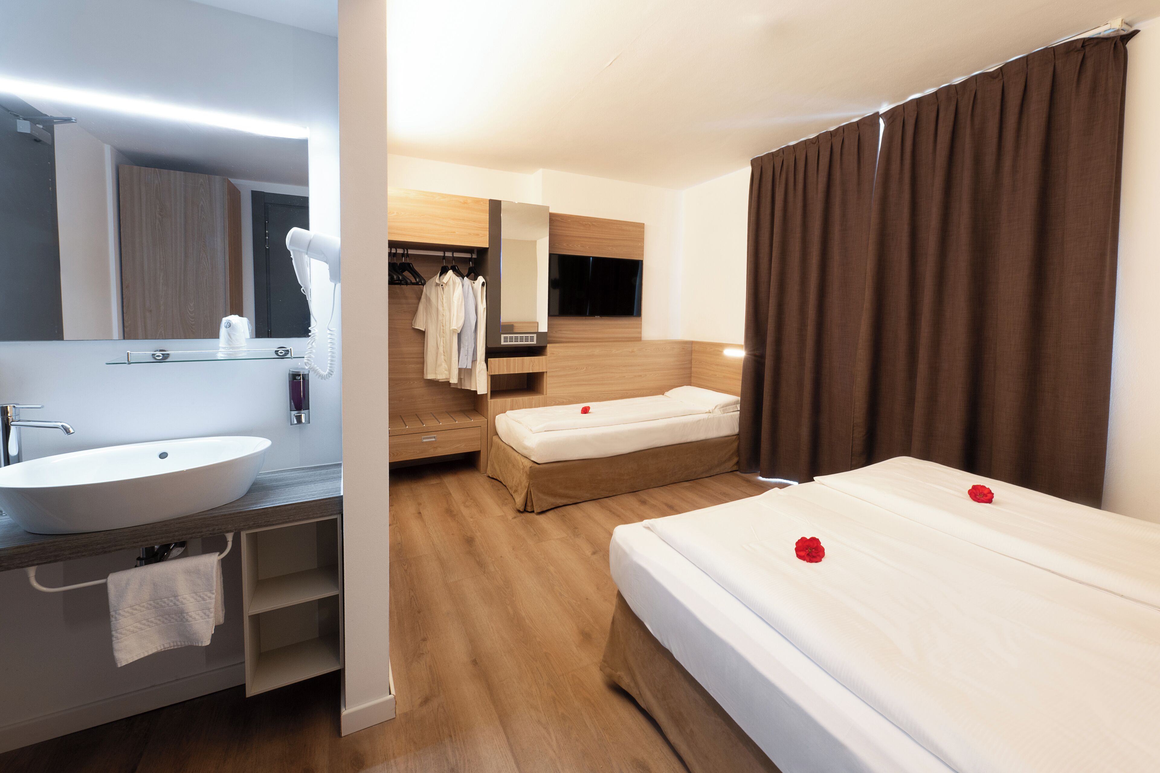 A hotel room with a large white sink, a mirror, a wooden vanity, a bed with white linens and red rose petals, a small bedside table, and a brown curtain.