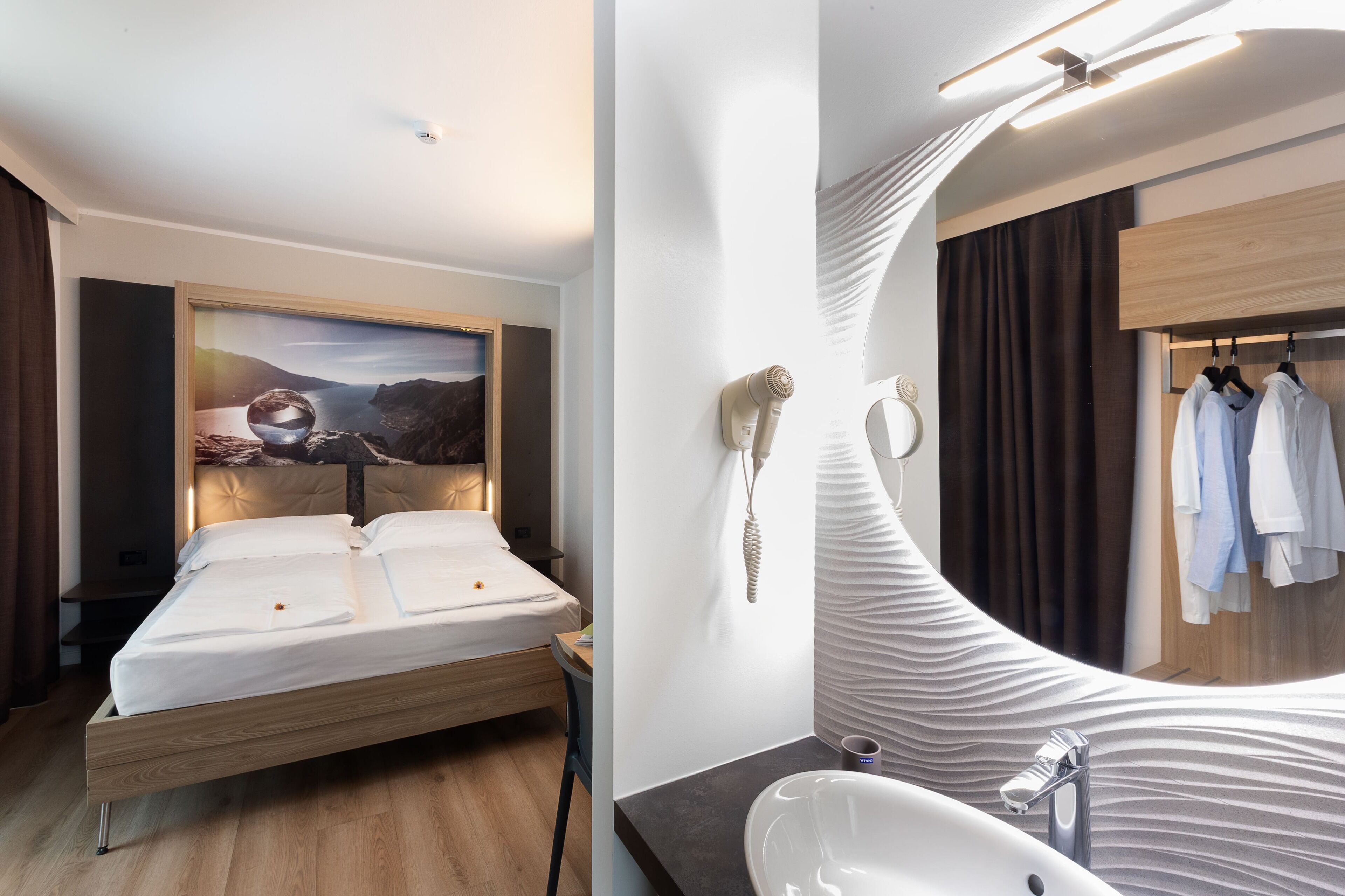 Una habitación de hotel moderna con una cama, un espejo grande, un tocador con lavamanos y un armario con ropa colgada.