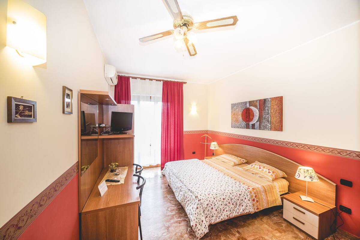 A hotel room with a bed, bedside table, desk, television, and a ceiling fan.