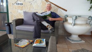 A man sitting on a wicker sofa reading a magazine in a room with a stone wall and a white ceramic pot. - Aurora Rooms (Lecce)