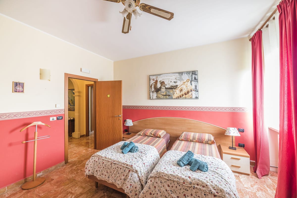 A room with two beds, a ceiling fan, a painting on the wall, and a red curtain.