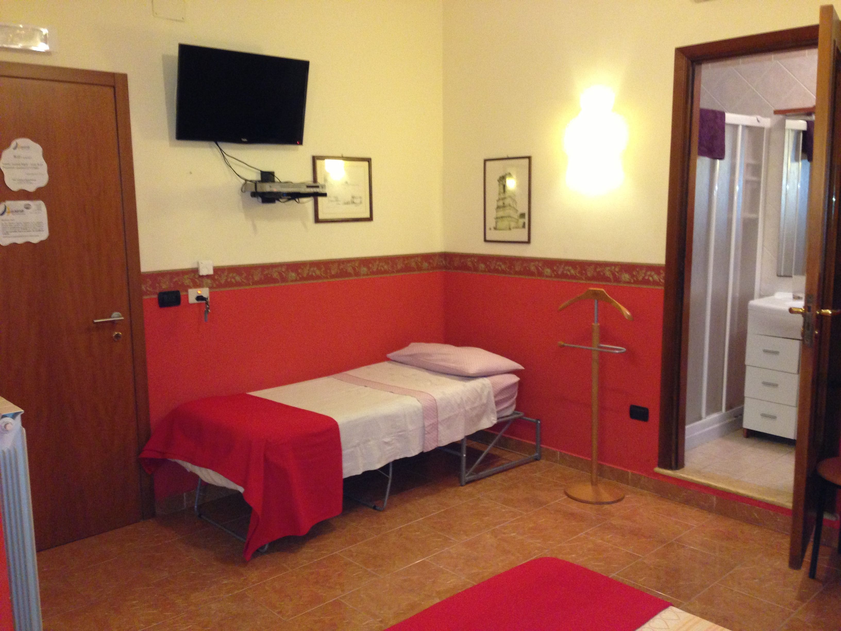 A small, single-bed room with a red and white color scheme, a television mounted on the wall, and a bathroom visible through an open door.