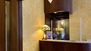 A reception desk with a wooden finish, a stained glass lamp, and a sign advising to maintain a minimum distance of 1 meter. - Hotel Bonazzi (Perugia)
