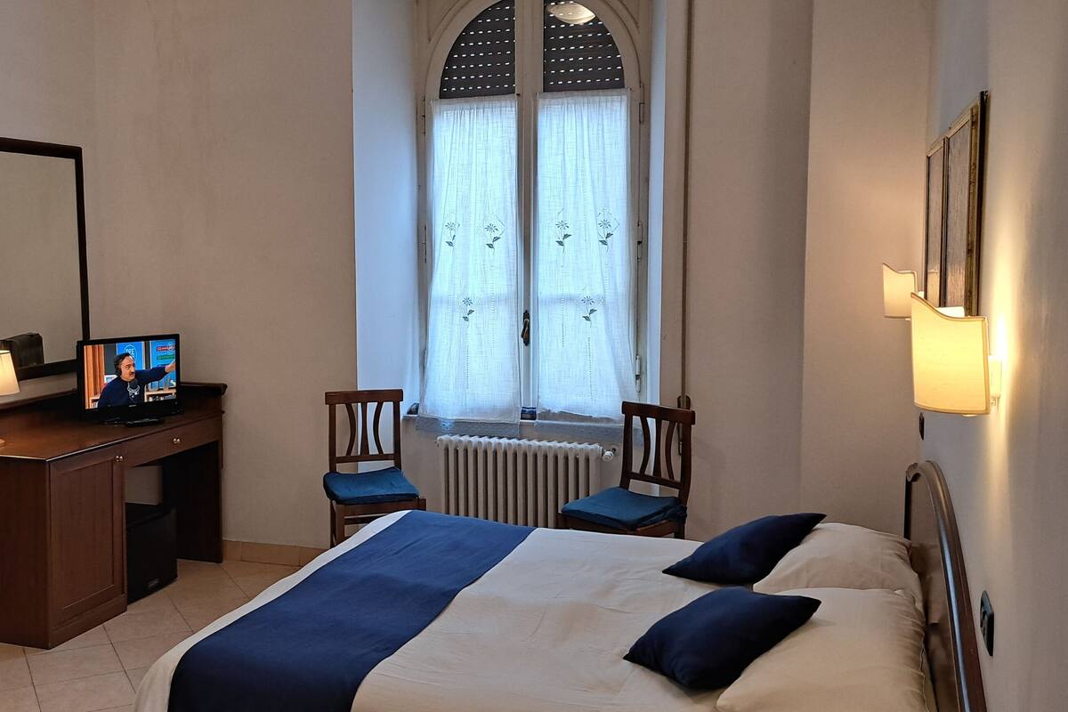Una habitación de hotel con cama, escritorio con computadora, silla, armario y ventana con cortinas.